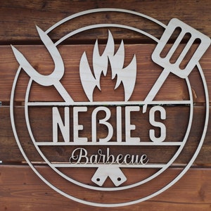 May include: A wooden sign with a circle design featuring a stylized flame, a spatula, and a fork. The sign reads "Nebie's Barbecue".