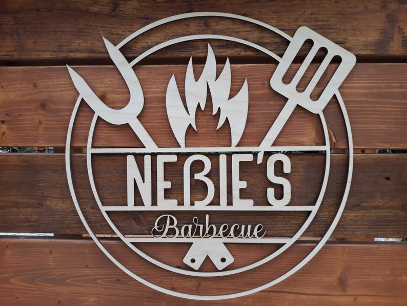 May include: A wooden sign with a circle design featuring a stylized flame, a spatula, and a fork. The sign reads "Nebie's Barbecue".