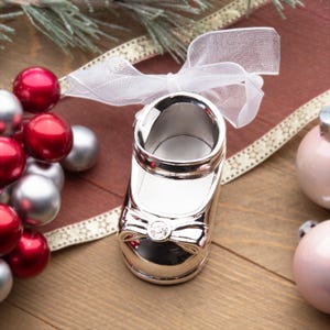 May include: Silver baby shoe ornament with a white bow and a clear rhinestone in the center. The ornament is hanging from a white ribbon.