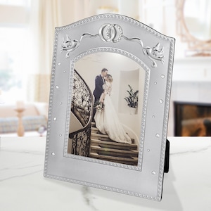 May include: Silver picture frame with a bevelled edge and a raised design of two doves and wedding rings. The frame has a white background and a clear opening for a photo.