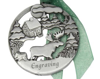 May include: A silver pewter Christmas ornament featuring a moose, a cabin, and pine trees. The ornament has a green ribbon and the word "Engraving" on the bottom.