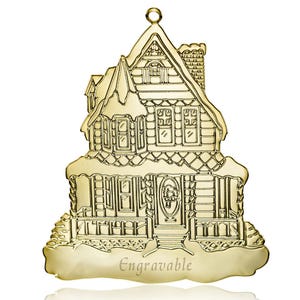 Peut inclure: Ornement en métal doré en forme de maison avec de la neige sur le toit. La maison a des fenêtres et une porte. L'ornement est gravable.