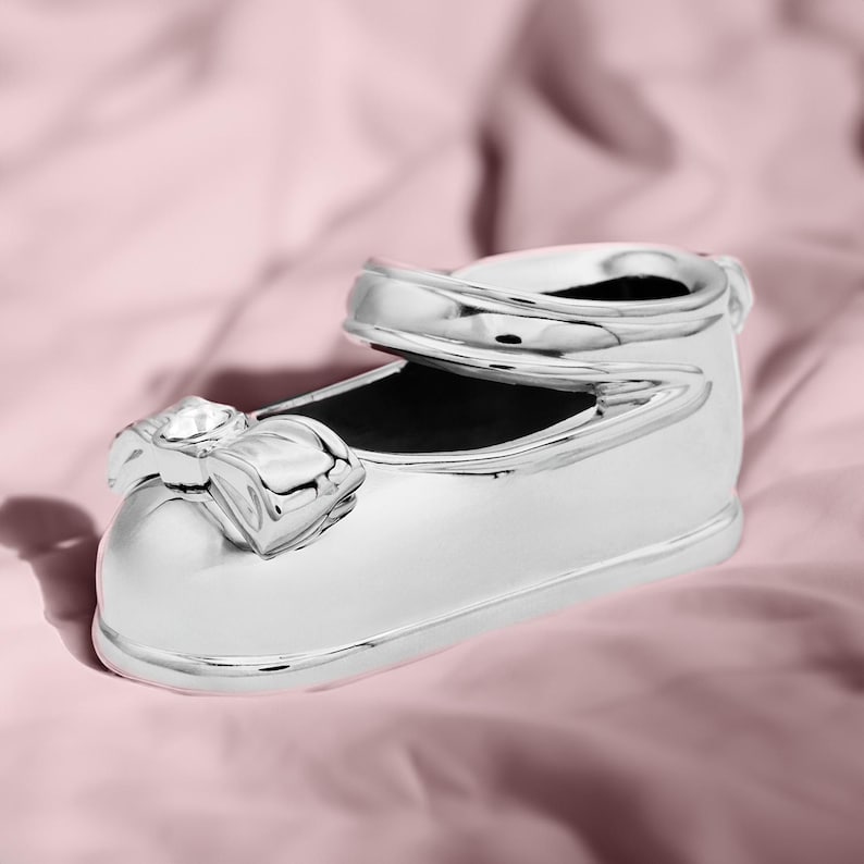 May include: A silver baby shoe with a bow and a crystal on top. The shoe is on a pink fabric background.
