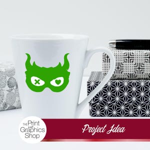 May include: White ceramic mug with a green superhero mask design. The mask has horns, an X for one eye, and a heart for the other. The mug is next to patterned boxes and a pen. The text "Project Idea" is on a red banner.
