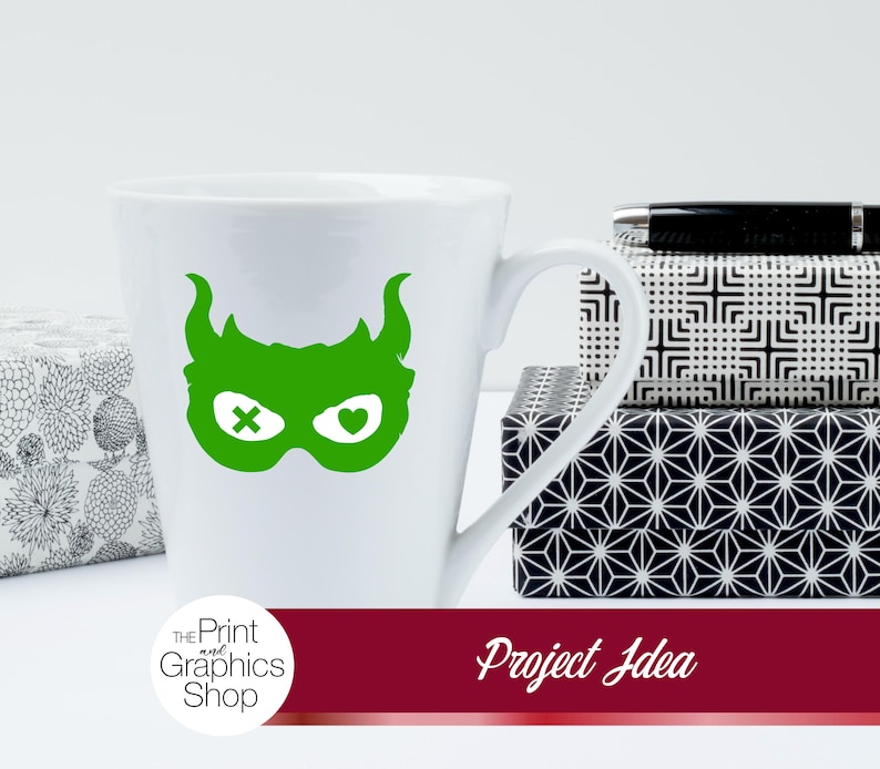 May include: White ceramic mug with a green superhero mask design. The mask has horns, an X for one eye, and a heart for the other. The mug is next to patterned boxes and a pen. The text "Project Idea" is on a red banner.