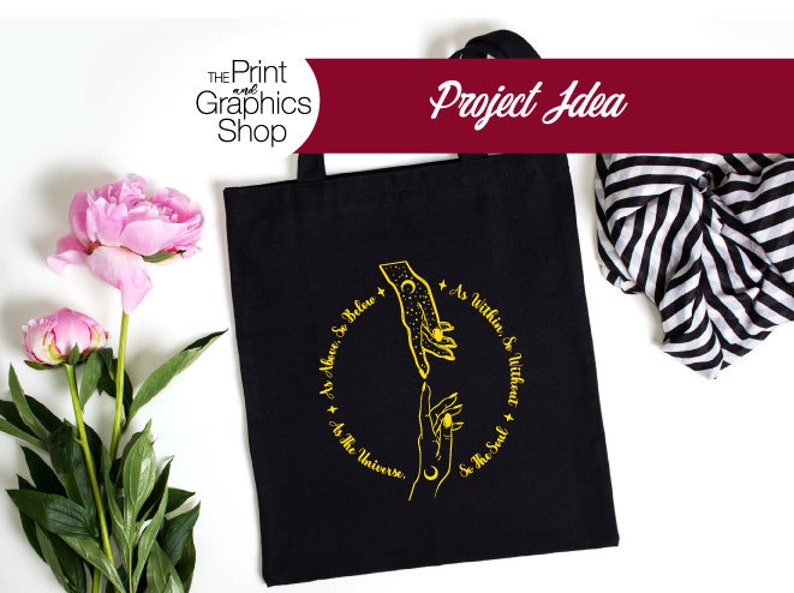 May include: Black tote bag with a gold design featuring two hands reaching towards each other, surrounded by text. The text reads "As Above, So Below, As Within, So Without, As The Universe, So The Soul." The bag has a handle and is on a white surface.