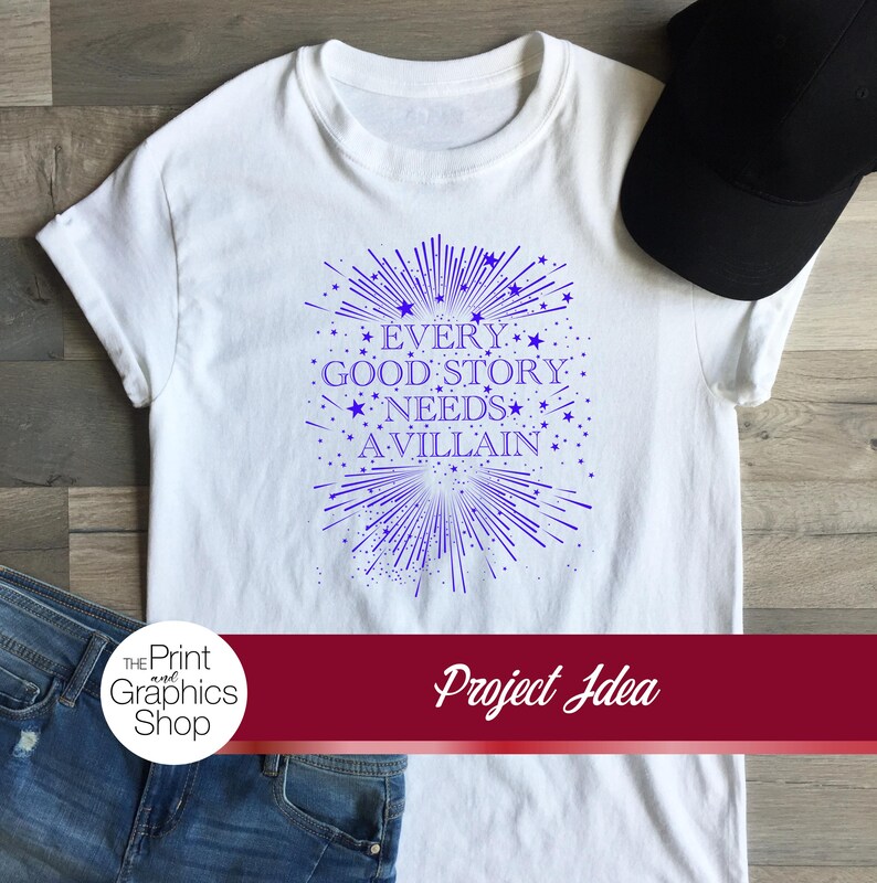 May include: A white short-sleeved t-shirt featuring a blue graphic print with the text "EVERY GOOD STORY NEEDS A VILLAIN" surrounded by radiating lines and small star shapes. The shirt is displayed alongside a black baseball cap and blue jeans.