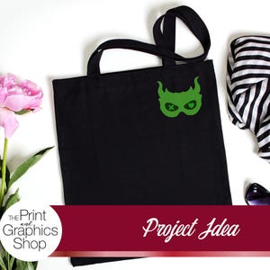May include: Black tote bag with a green graphic design, a black and white striped scarf, and pink flowers. The tote bag has a green mask design. The text "Project Idea" is on a red banner.