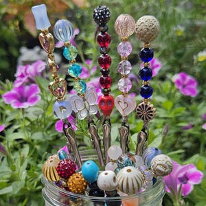 Peut inclure: Collection de pinces décoratives perlées dans un bocal en verre. Les pinces présentent divers motifs, notamment des cœurs, des crânes et des motifs floraux, avec des couleurs variées comme le rouge, le bleu et l'or. Le bocal est placé devant des fleurs et de la verdure violettes.