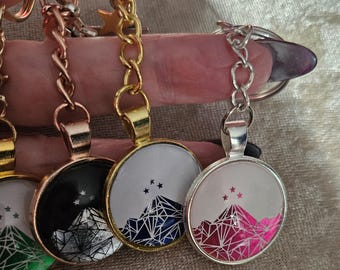 Könnte beinhalten: Eine Sammlung von vier runden Schlüsselanhängern mit einem geometrischen Bergdesign und drei kleinen Sternen über den Gipfeln. Die Anhänger sind in roségoldene, goldene und silberne Metallrahmen gefasst und zeigen jeweils ein farbiges Motiv.