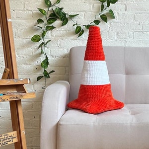 Puede incluir: Un cojín de cono de tráfico de punto rojo y blanco se encuentra en un sofá blanco. El sofá está frente a una pared de ladrillo blanco con una vid verde.
