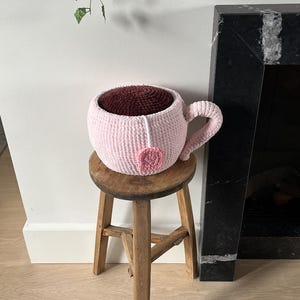 Puede incluir: Una gran taza de té rosa tejida a ganchillo, con asa y detalle de bolsita de té. El interior de la taza es de color burdeos oscuro, que se asemeja al té. La taza de té está sobre un pequeño taburete de madera rústica.