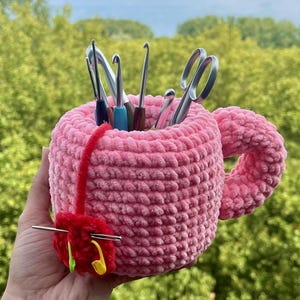 May include: A pink, plush, crocheted mug-shaped organiser filled with crochet hooks and scissors. A red yarn needle holder with a needle and stitch markers is attached. The background is a blurred view of green foliage.