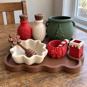 May include: A brown tray holds a red bottle with a snowflake, a white bottle with a snowman, a green cauldron with a tree, a white scalloped bowl with a gingerbread man stirrer, a red bowl with a bow and candy cane, and a red brick chimney.