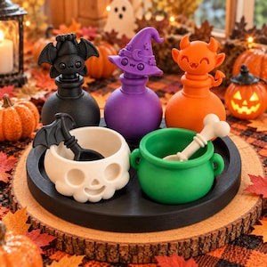 Może przedstawiać: Zestaw trzech butelek na mikstury w stylu Halloween w kolorach czarnym, fioletowym i pomarańczowym, z nakrętkami w kształcie nietoperza, kapelusza czarownicy i rogów diabła. Zawiera białą miskę w kształcie czaszki i zielony kociołek z łyżką.