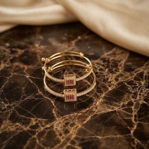 Può includere: Due braccialetti d'oro con elementi centrali quadrati color rubino. I braccialetti sono adornati con piccoli accenti scintillanti e sono impilati su una superficie di marmo. Lo sfondo presenta un tessuto dorato drappeggiato.
