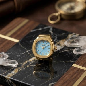 Puede incluir: Un reloj de anillo dorado con esfera azul claro y agujas doradas. El reloj tiene una esfera cuadrada con esquinas redondeadas. El reloj se exhibe sobre una superficie de mármol negro y dorado con cristales de cuarzo transparentes.