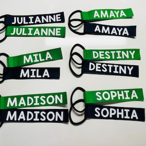 May include: Six pairs of personalized luggage tags with black elastic loops. Each tag has two pieces of fabric, one green and one navy blue, with white text. The names on the tags are Julianne, Amaya, Mila, Destiny, Madison, and Sophia.