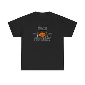 May include: Black t-shirt featuring a Halloween-themed design. The design includes the text "SALEM UNIVERSITY EST. 1692" with a jack-o'-lantern and candles. The shirt is made of a soft, comfortable material.