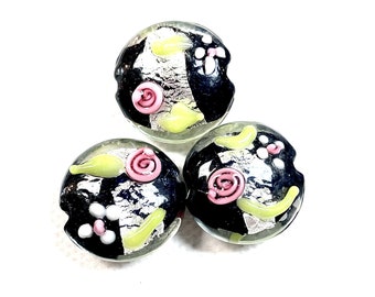 May include: Three round glass beads with black, white, silver, pink and yellow designs. The beads have a glossy finish.