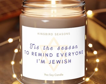 May include: A brown soy candle with a white label that reads "Tis the season to remind everyone I'm Jewish" and "9oz Soy Candle". The candle is lit and sitting on a wooden coaster.