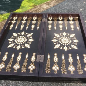 May include: A rectangular, open backgammon board featuring a dark brown wooden frame and a black interior surface. The board is decorated with intricate, light-colored floral mandala patterns in the center and ornate, elongated triangular point markers.