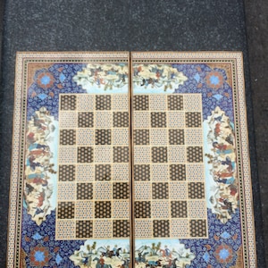 May include: A rectangular backgammon board featuring a central grid of alternating black and light-colored patterned squares. The border is decorated with intricate blue, gold, and white floral motifs and detailed miniature scenes of figures on horseback.