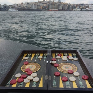 Könnte beinhalten: Ein Backgammon-Brett mit roten und weißen Spielsteinen und Würfeln ist im Freien aufgebaut. Das Brett ist offen, mit einem dunklen Holzrahmen und dekorativen kreisförmigen Mustern. Der Hintergrund zeigt eine Stadtlandschaft und Wasser.