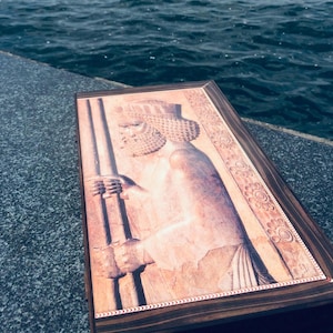 Könnte beinhalten: Eine rechteckige Holzschatulle mit einer detaillierten, hellen Reliefschnitzerei einer antiken persischen Figur auf dem Deckel. Die Figur trägt eine traditionelle Kopfbedeckung und hält zwei lange Stäbe. Der Rahmen besteht aus dunklem Holz.
