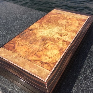 Könnte beinhalten: Eine rechteckige Holzkiste mit einem Vintage-Kartenmuster auf dem Deckel. Die Kiste hat ein dunkles Holzmaserungsmuster mit glänzendem Finish. Die Karte zeigt eine Weltkarte mit komplizierten Details und einem cremefarbenen Hintergrund.