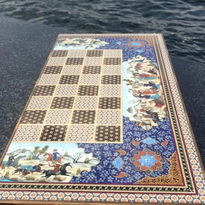 May include: A rectangular wooden game board featuring a central grid of alternating light and dark patterned squares. The border is decorated with intricate blue, gold, and white floral motifs and detailed miniature paintings of figures on horseback.