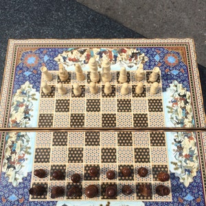 May include: A square chess board featuring a central grid of alternating black and beige patterned squares. The board is decorated with intricate, colorful miniature paintings of figures and horses. Light beige and dark brown wooden chess pieces.