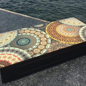 Könnte beinhalten: Eine rechteckige schwarze Schachtel mit einer Oberseite, die mit einem lebendigen, mehrfarbigen Mandalamuster verziert ist. Das Design besteht aus mehreren überlappenden kreisförmigen Motiven in Gelb-, Türkis-, Orange- und Dunkelblautönen.