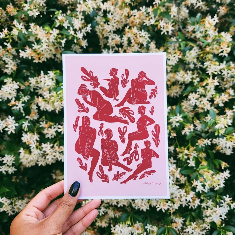 May include: A pink art print featuring red illustrations of figures in various poses, interspersed with leaf-like shapes. The print is held against a backdrop of white flowers and green foliage. The artist's signature is visible.