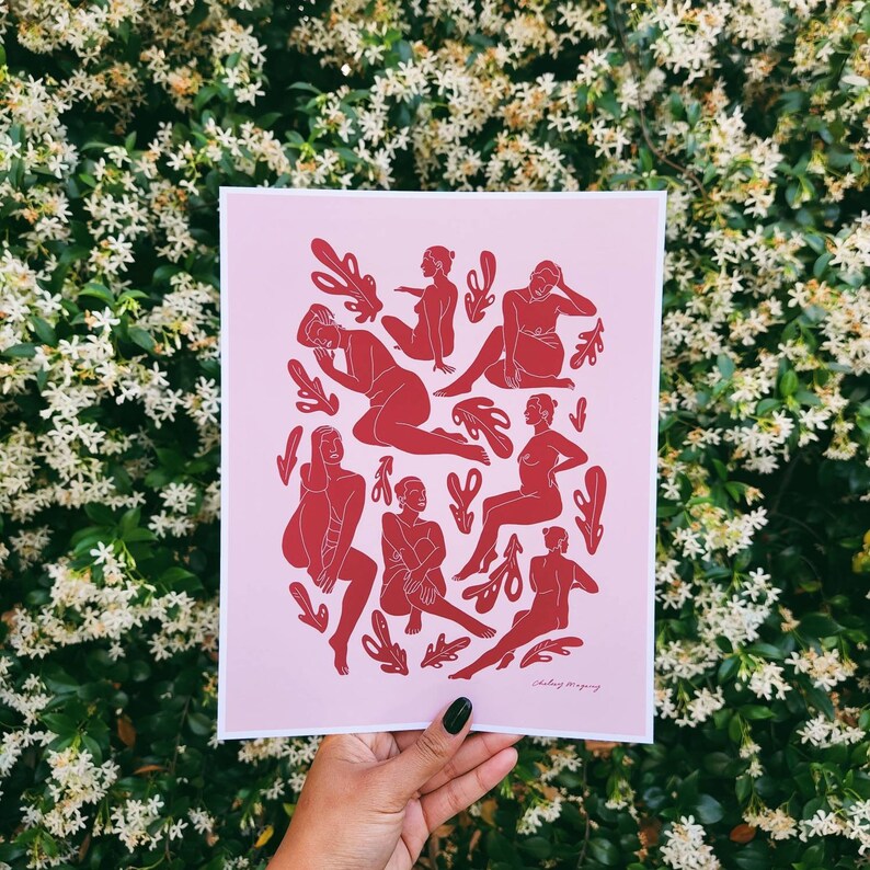 May include: A pink art print featuring red illustrations of figures in various poses, interspersed with leaf-like shapes. The artwork is held by a hand against a backdrop of white flowers.