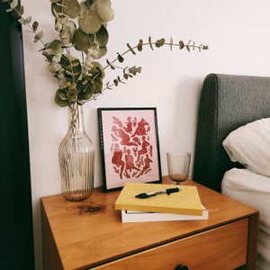May include: A wooden nightstand with a framed pink print, a clear glass vase with eucalyptus, a yellow notebook, a pen, and a clear glass. The print features red figures. The nightstand is next to a bed with a gray headboard.