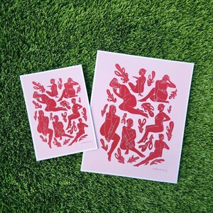 May include: Two art prints featuring red figures in various poses and leaf-like shapes on a light pink background. The prints are on white-bordered paper. The prints are displayed on a green grassy surface.