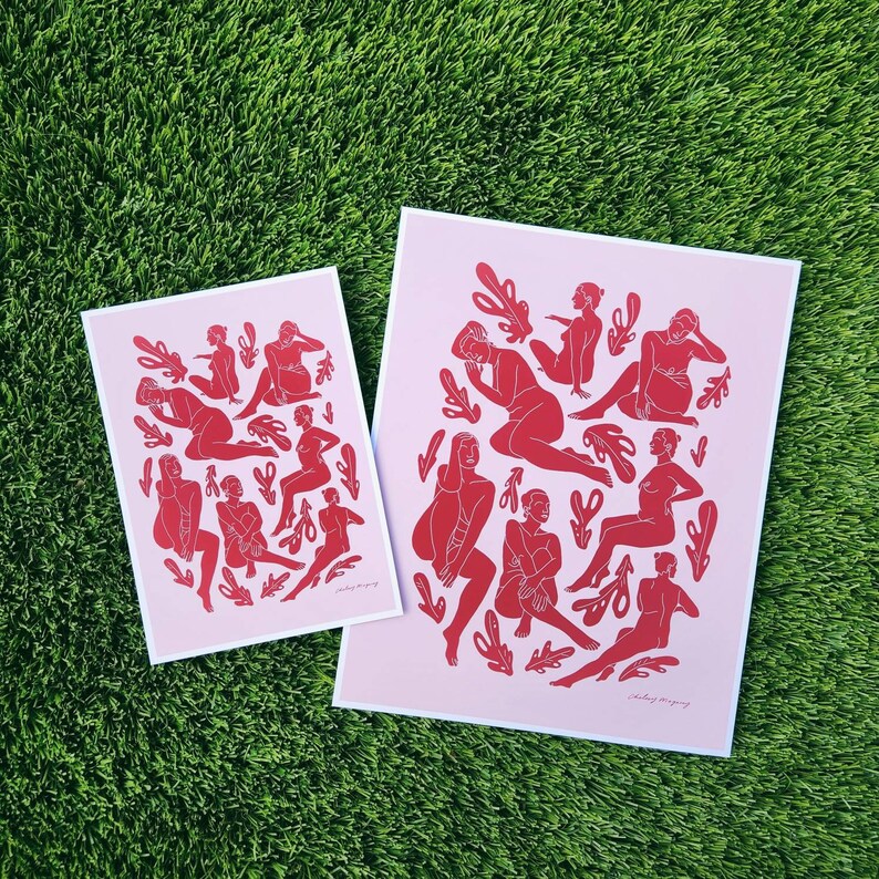 May include: Two art prints featuring red figures in various poses and leaf-like shapes on a light pink background. The prints are on white-bordered paper. The prints are displayed on a green grassy surface.