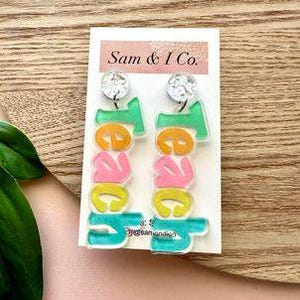 May include: A pair of colourful acrylic earrings with the word "Tees" spelled out in a playful font. The earrings are green, yellow, pink and orange.