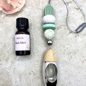 May include: A car air freshener with a wooden and silver diffuser, a green and white beaded chain, and a small bottle of essential oil labelled "Musk Stick".