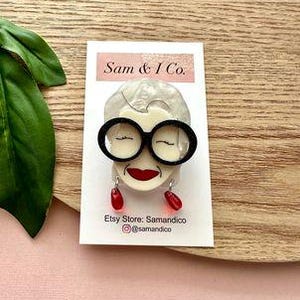 May include: White resin brooch featuring a black framed glasses and red lips. The brooch is shaped like a woman's face. The text on the card reads "Sam & I Co. Etsy Store: Samandico @samandico"
