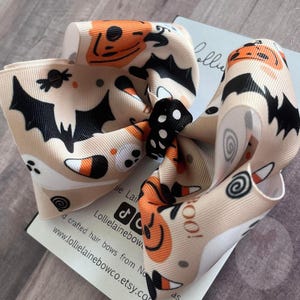 May include: A beige Halloween hair bow with a repeating pattern of pumpkins, bats, ghosts, and candy corn. The bow has a black centre and is likely made of a satin-like material. A festive accessory for the spooky season.