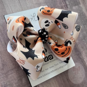 May include: A large Halloween-themed hair bow with a tan background, orange pumpkins, black bats, and white ghosts. The bow has a black and white polka dot centre and the word "BOO" printed on the ribbon.