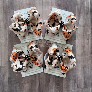 May include: Four Halloween hair bows. Each bow has a beige background with orange pumpkins, black bats, white ghosts, and candy corn. The bows are tied with a black and white polka dot centre.