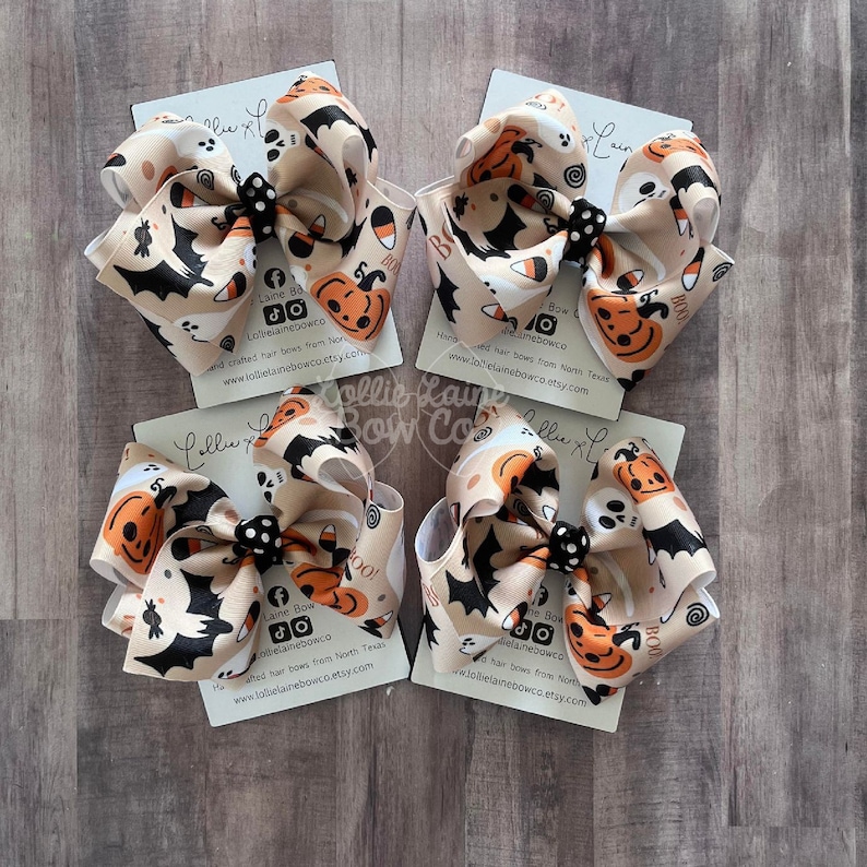 May include: Four Halloween hair bows. Each bow has a beige background with orange pumpkins, black bats, white ghosts, and candy corn. The bows are tied with a black and white polka dot centre.