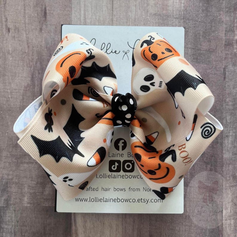May include: A beige Halloween hair bow with orange, black, and white patterns. The bow features bats, ghosts, and jack-o'-lanterns. The centre is a black bow with white polka dots. The word "BOO!" is visible.