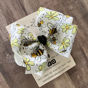 May include: A white hair bow with a yellow and black bee and daisy print. The bow is tied with a black elastic band. The bow is on a white card with the text "Lollie Bow Co" and "Hand crafted hair bows from North Texas www.lollielainebowco.etsy.com".