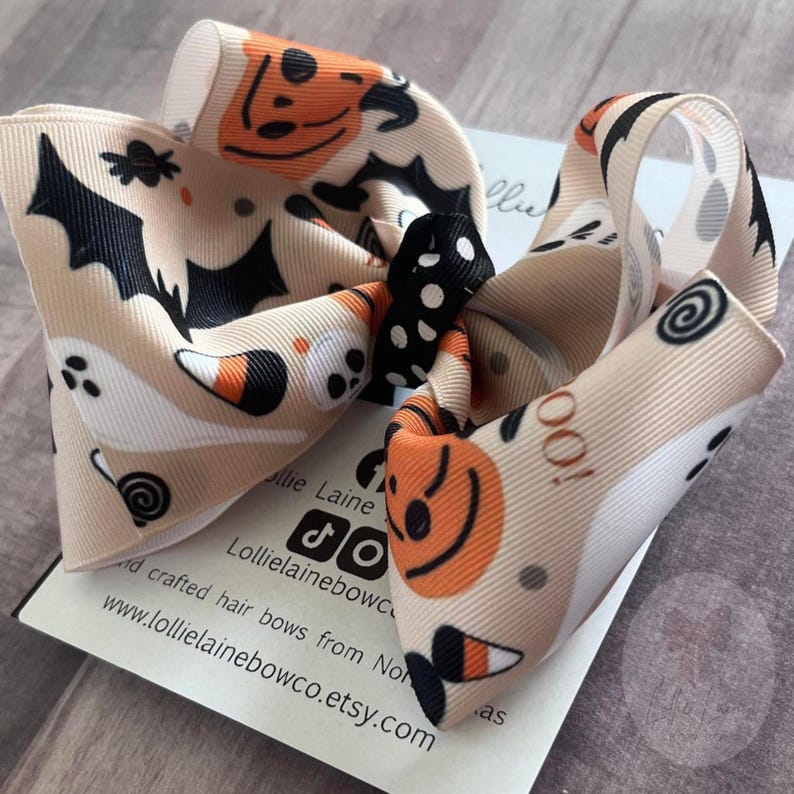 May include: A beige Halloween hair bow with orange pumpkins, black bats, white ghosts, and candy corn. The bow has a black and white polka dot centre and is printed with the text "Lollie Laine Bow Co."