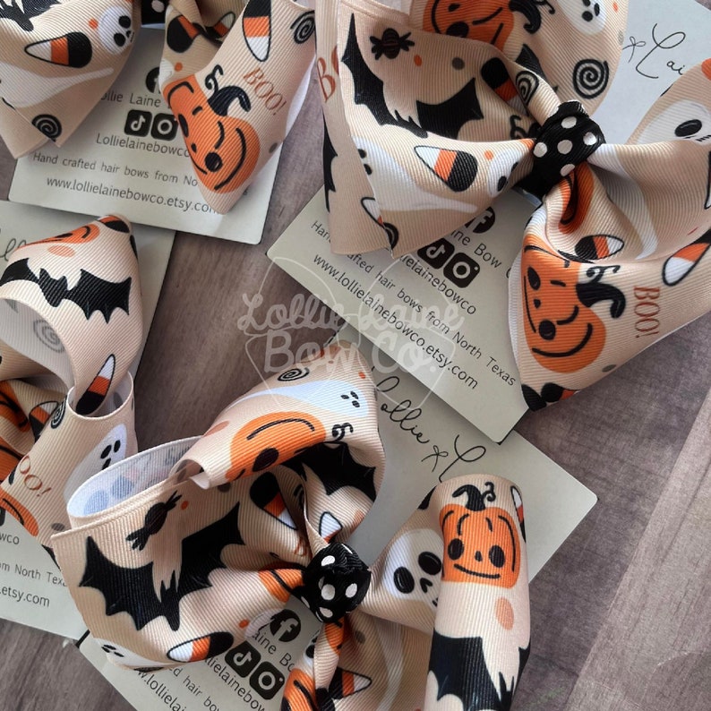 May include: A collection of Halloween hair bows with a beige background. The bows display repeating patterns of pumpkins, ghosts, bats, and candy corn. Each bow has a black and white polka dot centre. The word "BOO!" is printed on the bows.