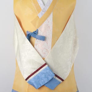 Może przedstawiać: Tradycyjny koreański hanbok w kolorze żółtym i białym z niebieskimi i bordowymi wykończeniami. Hanbok ma biały panel z koronki i niebieską wstążkę zawiązaną z przodu.